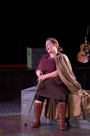 Giant-bosomed Jill Falstaff in red knit blouse, brown skirt with black leggings underneath, brown boots, and brown cloak, sits with a cain in her hand. 